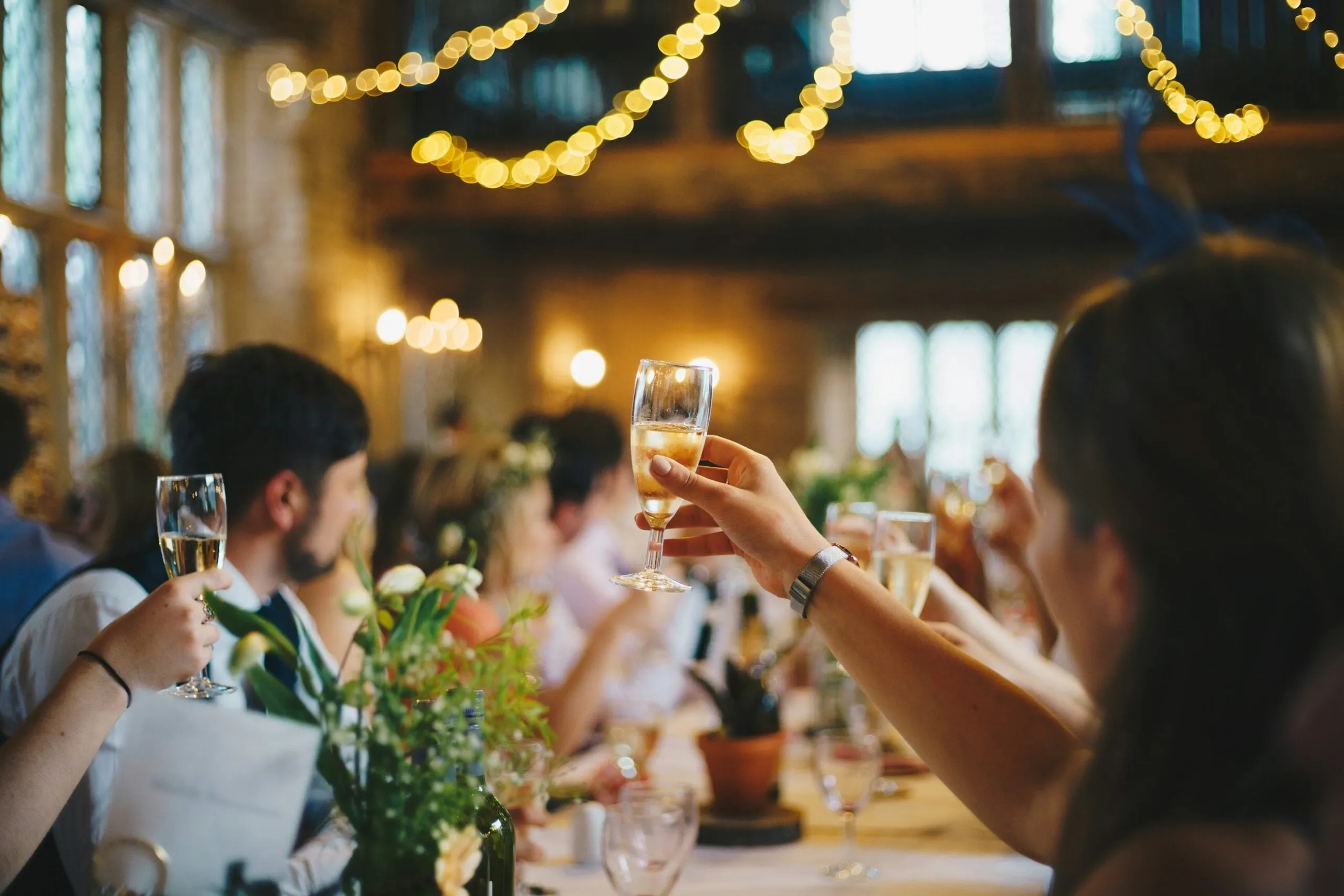 Invités levant leur verre autour d’une longue table dressée, sous des guirlandes lumineuses